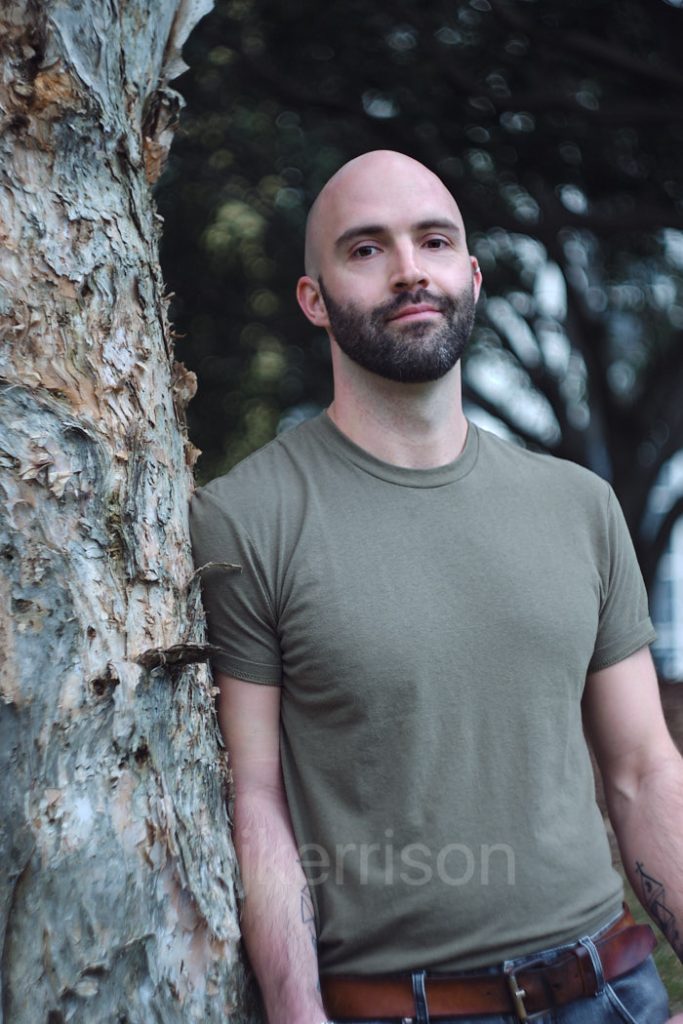 Pierre wears a military green T shjirt and rests against a paperbark tree. He looks to camera. 