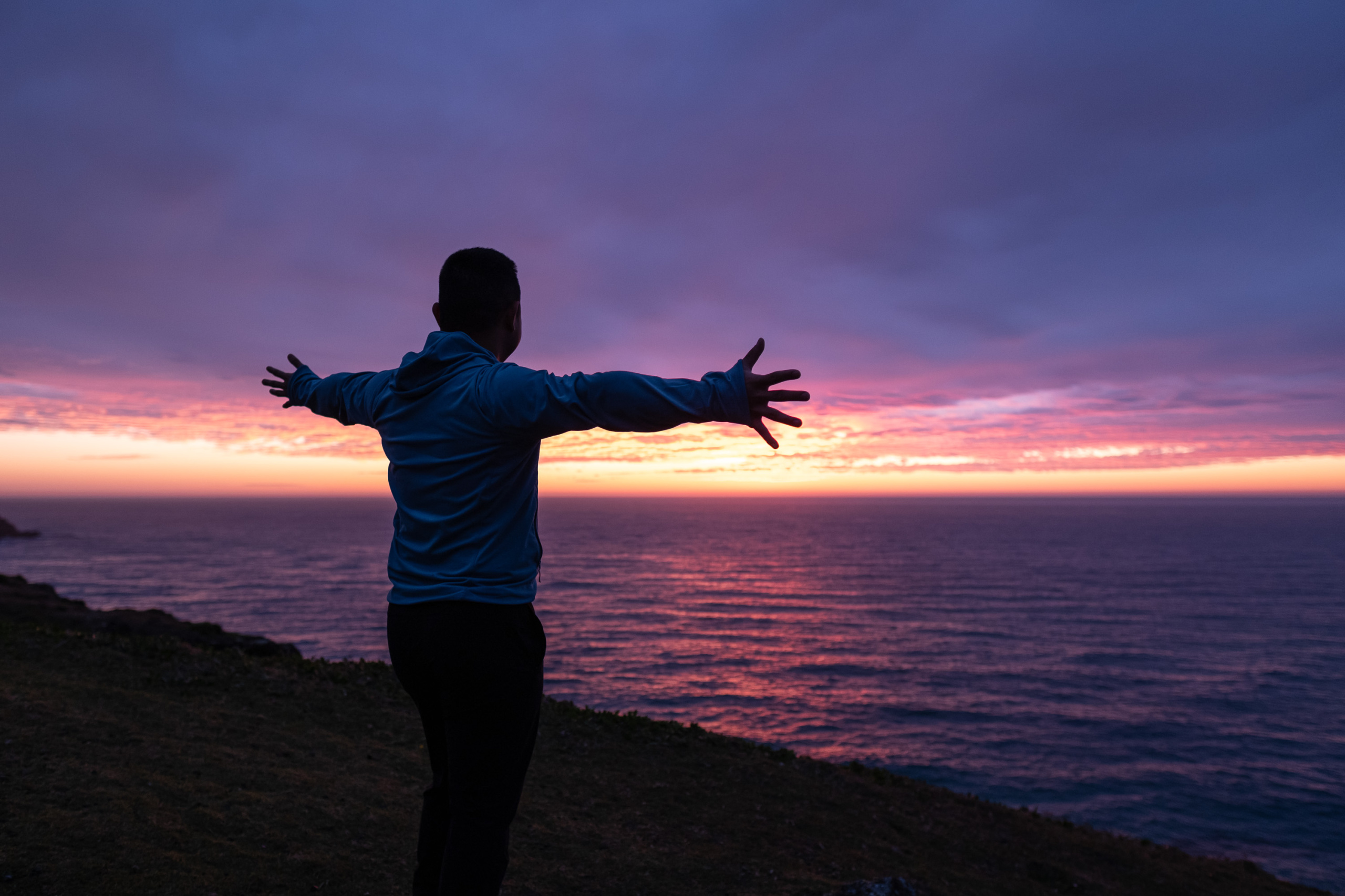 A man stands on a hillside with his arms stretched open. He has his back to camera as the sun comes up over the ocean. 